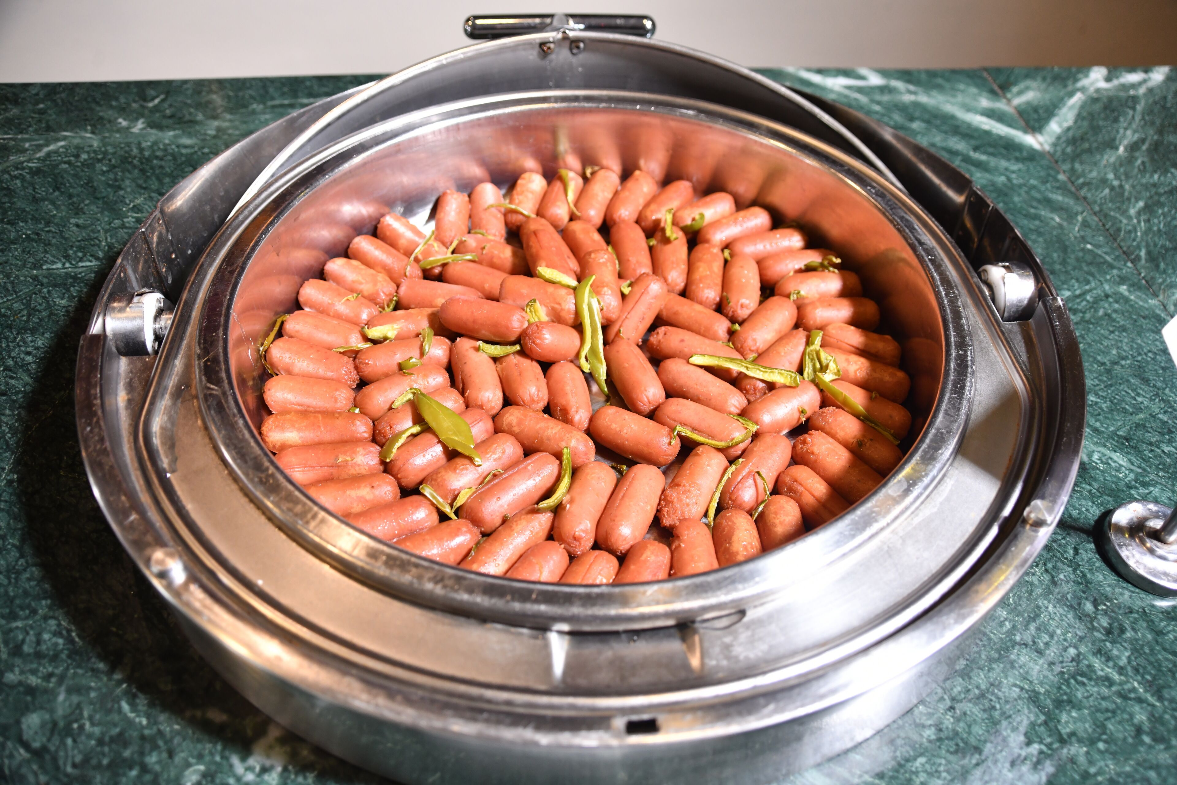A round, metallic container filled with small, round, orange objects, likely food items, on a green marble surface.