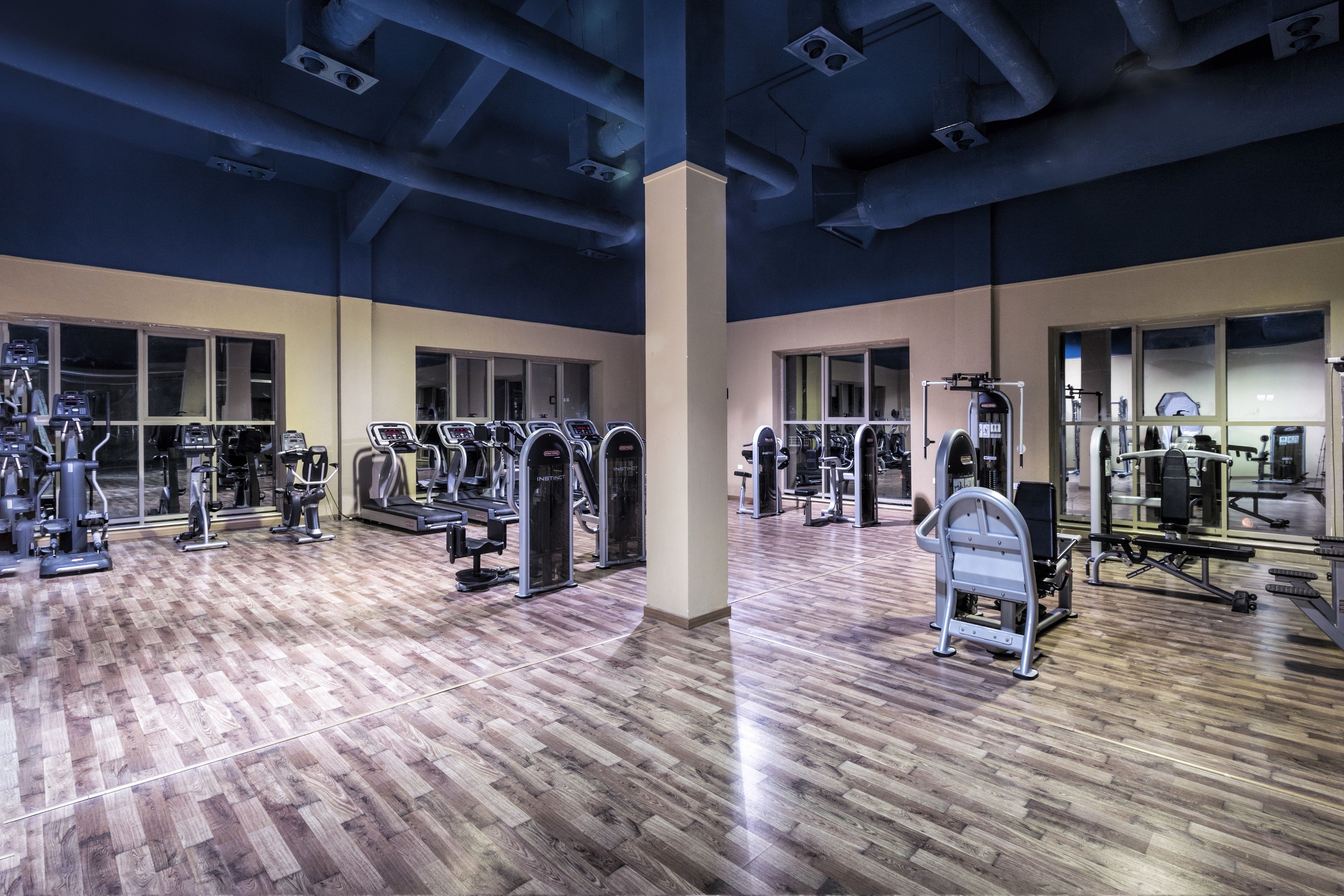 A gym with various exercise machines, wooden flooring, and large windows.