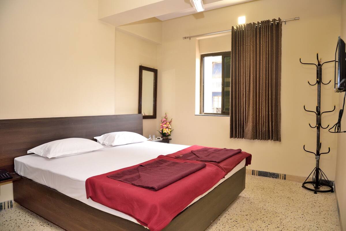 A neatly made bed with white linens and a red blanket, a wooden headboard, a small bedside table with a phone, a window with curtains, and a coat rack.