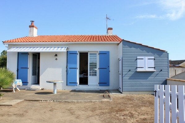 A two-story house with a red-tiled roof, blue shutters, and a small patio area with a table and chairs.