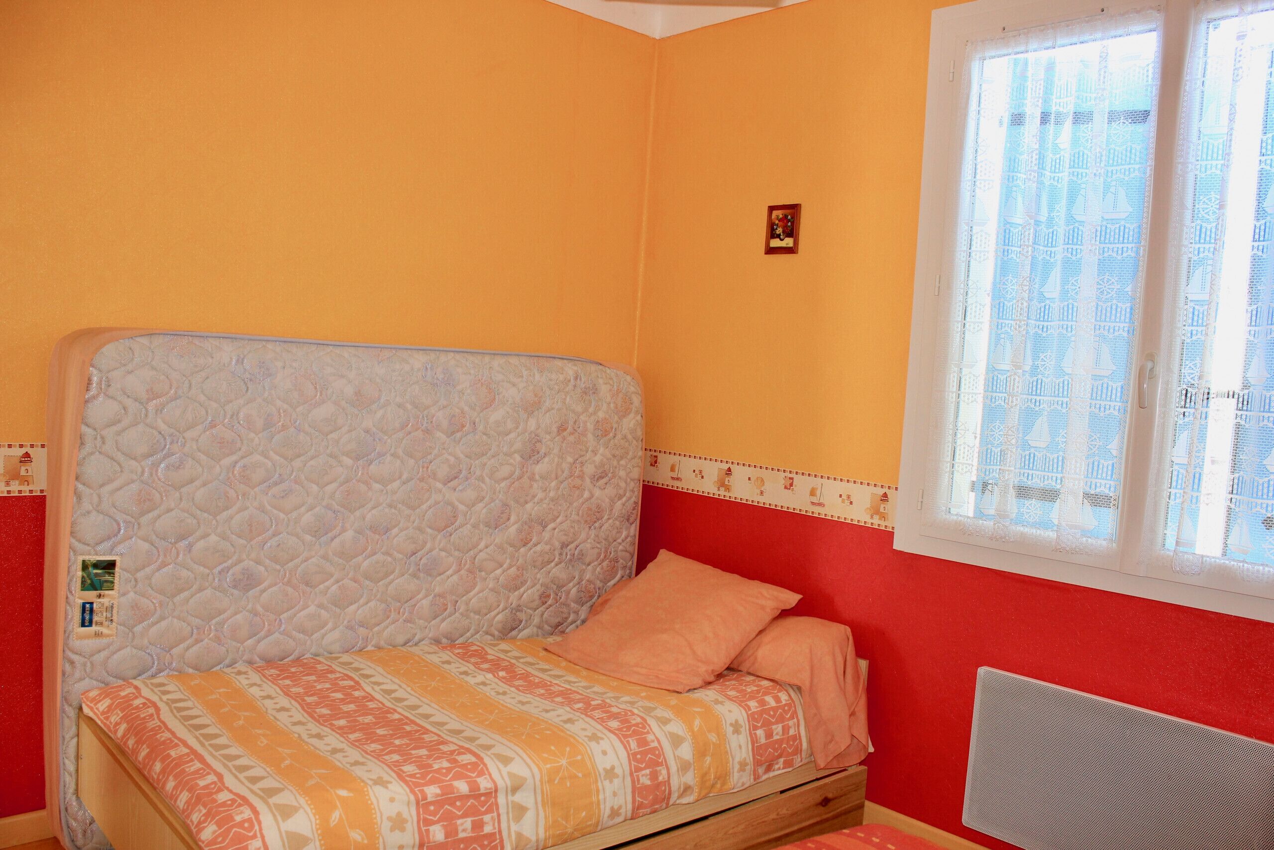A single bed with a patterned bedspread in a room with orange walls and a window with lace curtains.