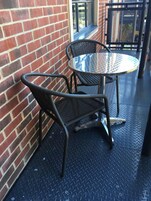 Una mesa metálica redonda con superficie de vidrio, acompañada por dos sillas de mimbre, ubicada frente a un muro de ladrillo con una barandilla metálica al fondo.