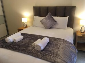 A neatly made bed with a grey textured bedspread, two rolled white towels, and a single grey pillow. - Blue sky at Wallis square (Farnborough)