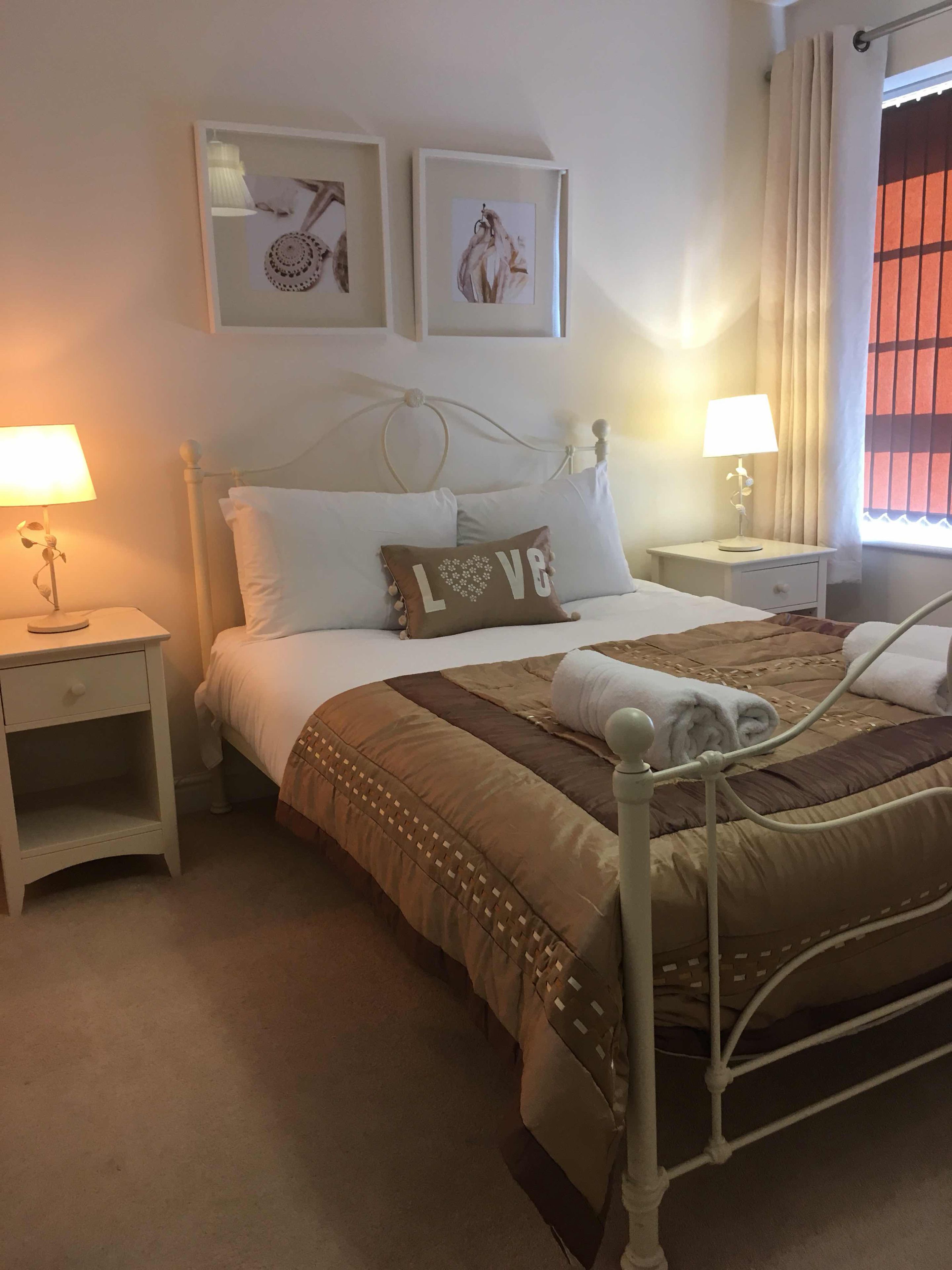 A neatly arranged bedroom with a bed, two bedside tables, a nightstand, a window with curtains, and two wall-mounted artworks.
