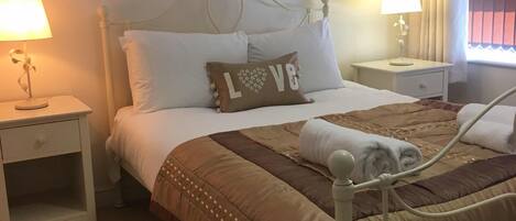 A neatly arranged bedroom with a bed, two bedside tables, a nightstand, a window with curtains, and two wall-mounted artworks.