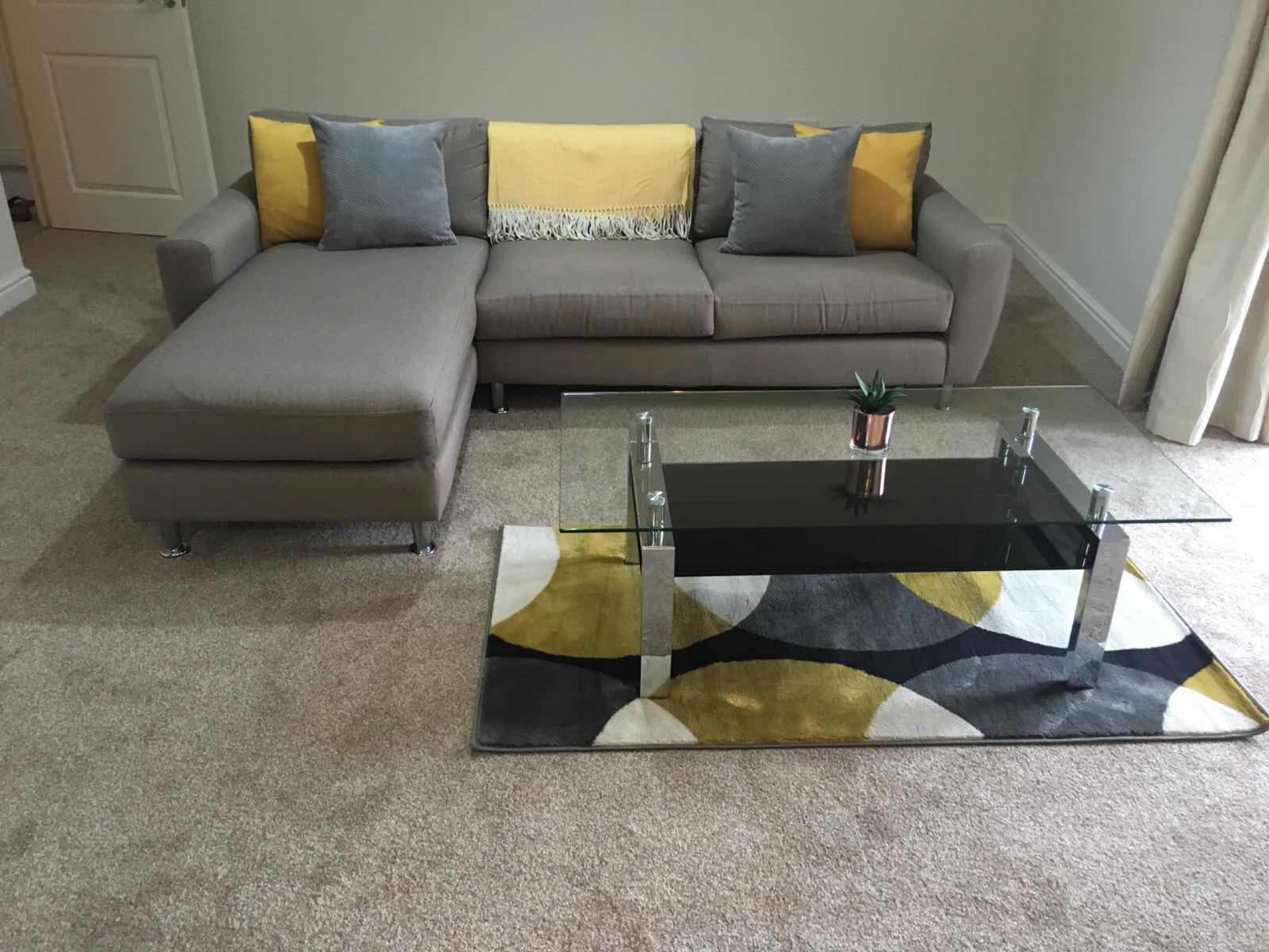 Ein Wohnzimmer mit einem grauen Ecksofa, einem Glastisch und einem gemusterten Teppich.