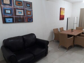 A living room with a black leather sofa, a wooden dining table with four chairs, a refrigerator, and a wall-mounted abstract painting. - Casa VicBec (Playa del Carmen)