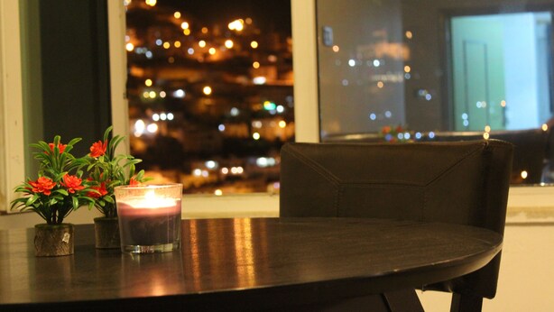 Una mesa con una vela y un florero, una silla y vista de un paisaje urbano de noche.