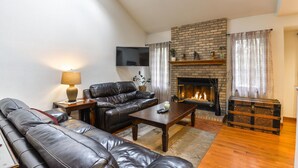 A living room with a fireplace, leather sofas, a coffee table, and a TV mounted on the wall. - 5 Bedroom house with a Hot Tub and Home Theater! (Fort Collins)