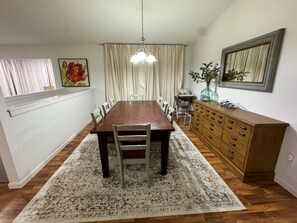 A dining room with a wooden table, white chairs, a patterned rug, a wooden cabinet, and a hanging light fixture.