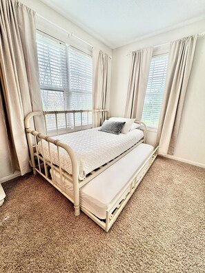 A single-bed room with a white metal frame, beige curtains, and a carpeted floor.