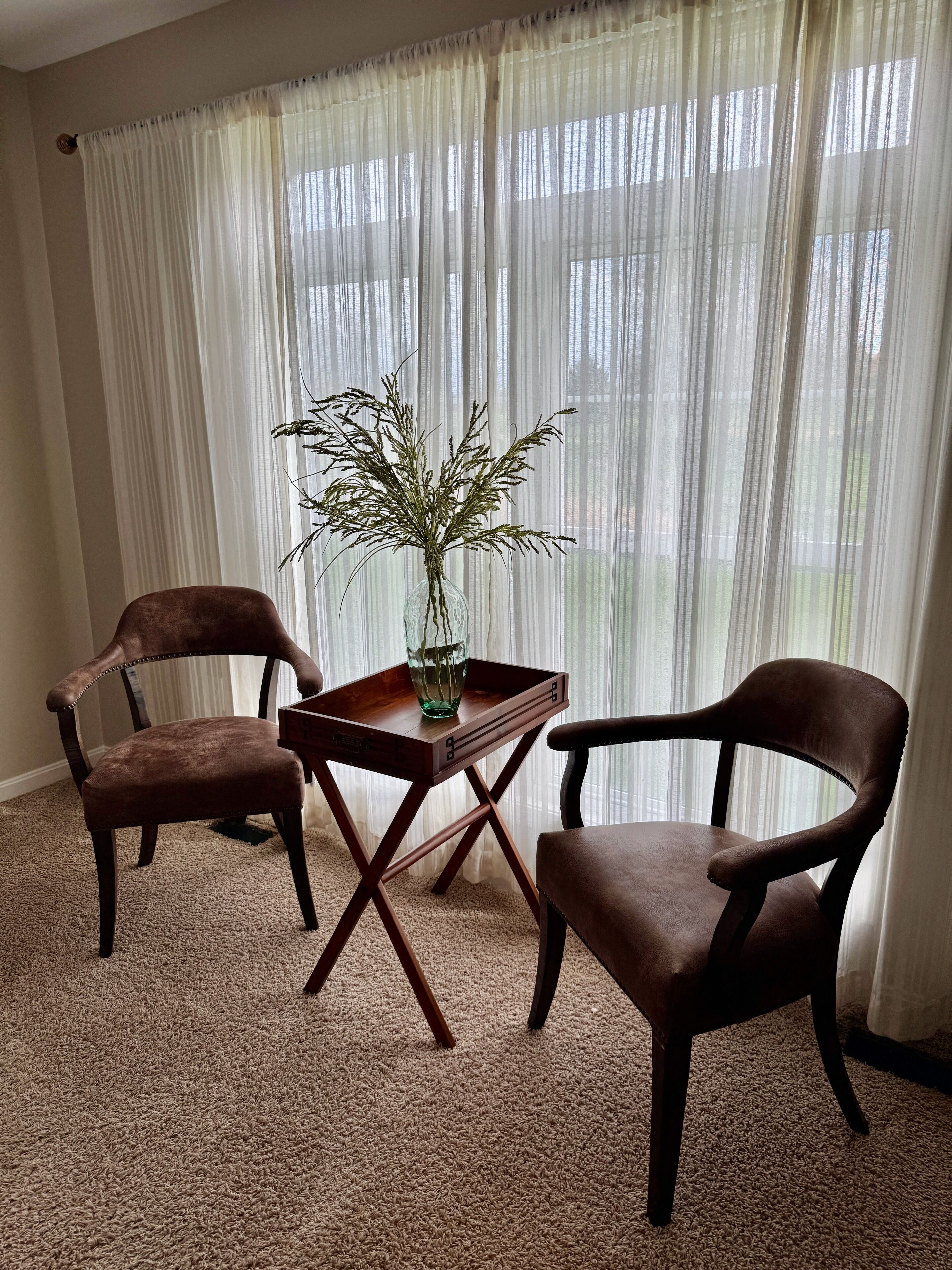 Une pièce avec deux fauteuils en cuir marron, une petite table en bois ornée d’un vase contenant des plantes séchées et de fines rideaux blancs.