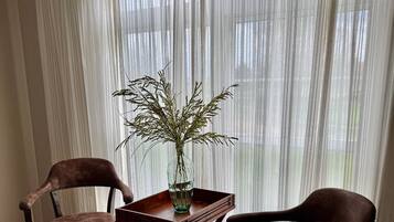 Une pièce avec deux fauteuils en cuir marron, une petite table en bois ornée d’un vase contenant des plantes séchées et de fines rideaux blancs.