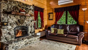 Ein gemütliches Blockhauskaminzimmer mit Kamin aus Stein, Ledersofa und Blick auf den Wald durch große Fenster.