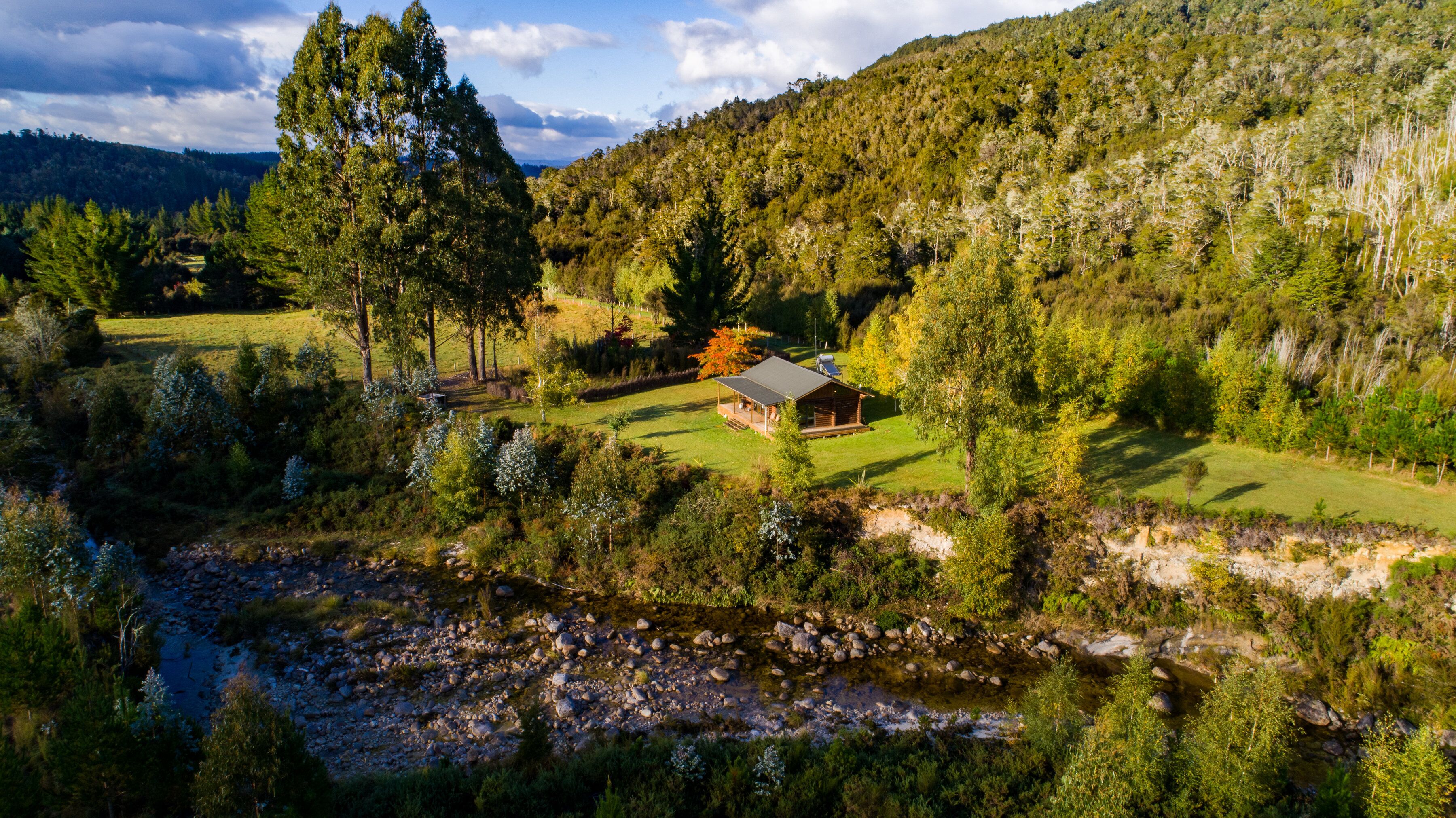 A cabin surrounded by a forested landscape with a river in the foreground.