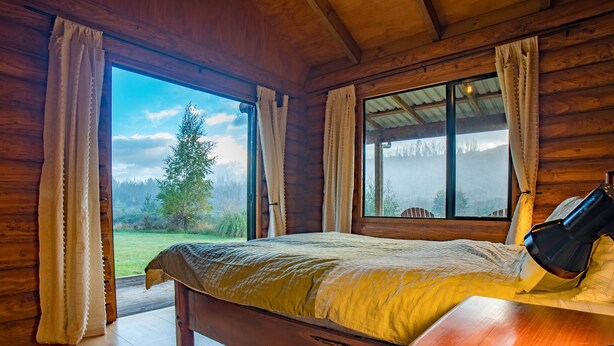 A wooden cabin bedroom with a bed, a nightstand, and a window with curtains. - Triple Tui - Log cabin, self contained, river side (Murchison)
