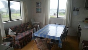 Een zithoek met een tafel bedekt met een blauw-wit geruit tafelkleed, omringd door stoelen. Er staat een bank met een bloemenmotief en een stoel met een gestreept kussen. De kamer heeft grote ramen met gordijnen en uitzicht op buiten.