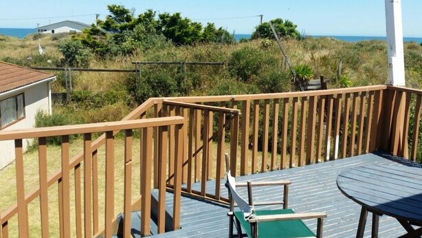 Een houten terras met een tafel en stoelen, met uitzicht op een gazon en de zee, met een gebouw op de achtergrond.