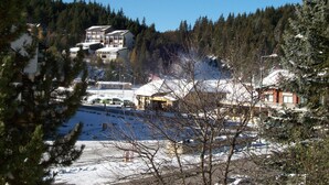 Un paysage enneigé avec des bâtiments, dont une structure à plusieurs étages dotée de balcons, le tout entouré d’une forêt dense.