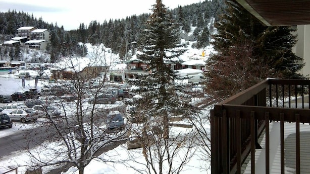 Une vue d’un parking avec plusieurs voitures, d’un immeuble doté d’un balcon et d’arbres couverts de neige.