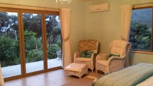 Ein Schlafzimmer mit einem Bett, zwei Korbstühlen, einem kleinen Tisch und Blick auf Grün hinter großen Schiebetüren.