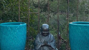 Eine Statue einer sitzenden Figur in meditativer Haltung, begleitet von zwei großen, blauen Pflanzkübeln, vor dem Hintergrund eines Metallgitters mit Blick auf einen Sonnenuntergang.
