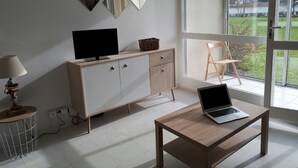 Ein modernes Zimmer mit Fernseher auf einer Kommode, einem Laptop auf einem Tisch und einem Stuhl neben einem großen Fenster.