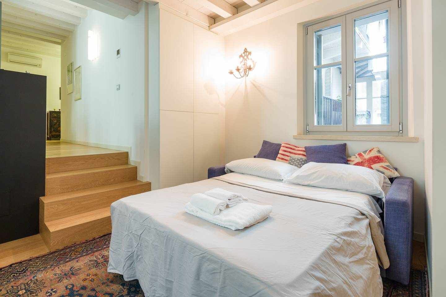 A bedroom with a bed, a window, a staircase, and a wall-mounted light fixture.