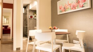 A dining area with a round table, white chairs, and a fruit bowl. There is a painting on the wall and a red couch in the background.