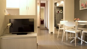 A modern living room with a flat-screen TV, a dining area with white chairs, and a kitchen visible in the background.