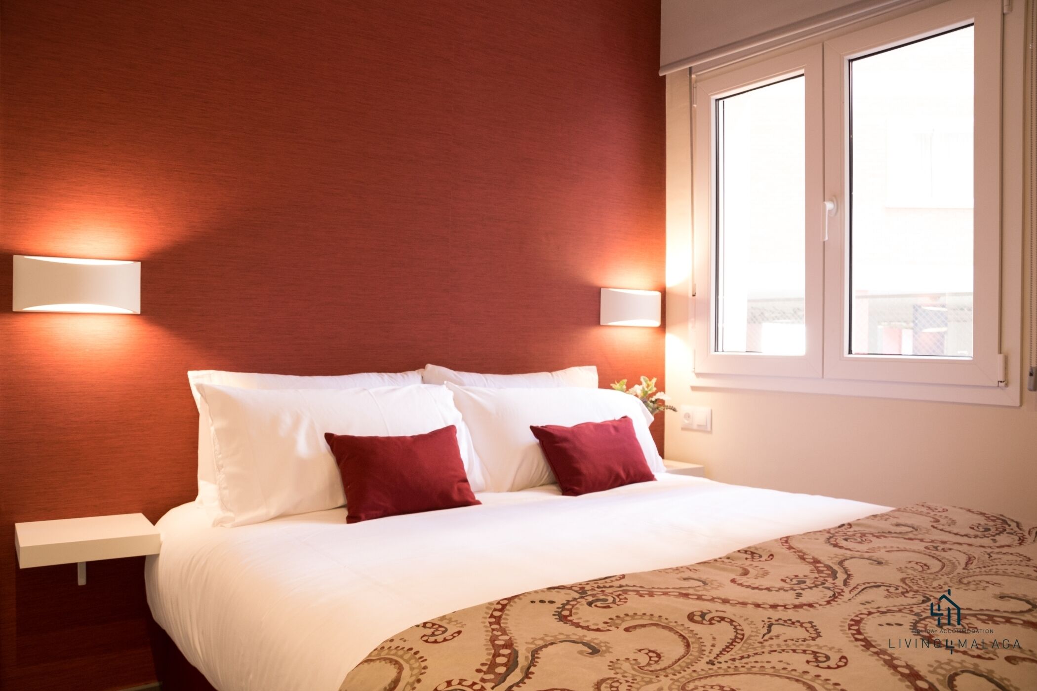 Ein Hotelzimmer mit einem Bett, weißer Bettwäsche, zwei roten Kissen, einer gemusterten Decke und zwei Wandleuchten.