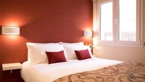 A hotel room with a bed, white bedding, two red pillows, a patterned throw, and two wall-mounted lights.