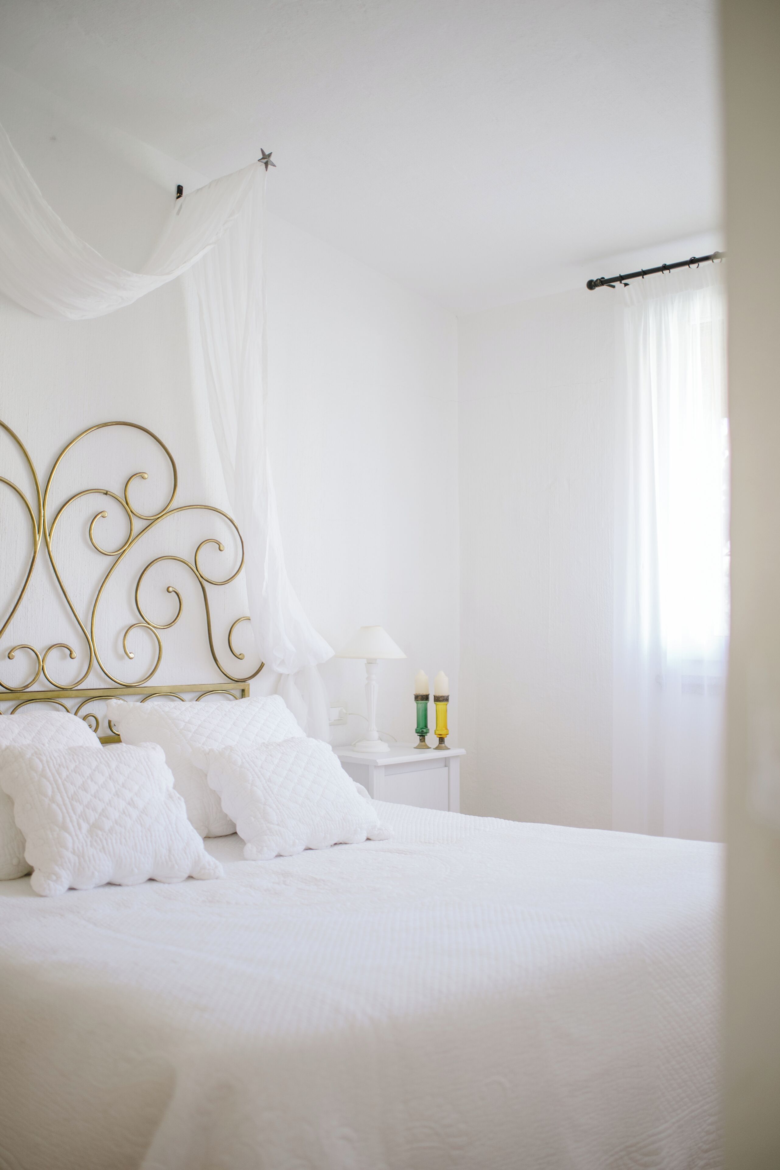 Una cama bien tendida con ropa blanca, cabecera de metal dorado y mesita de noche blanca con dos velas.