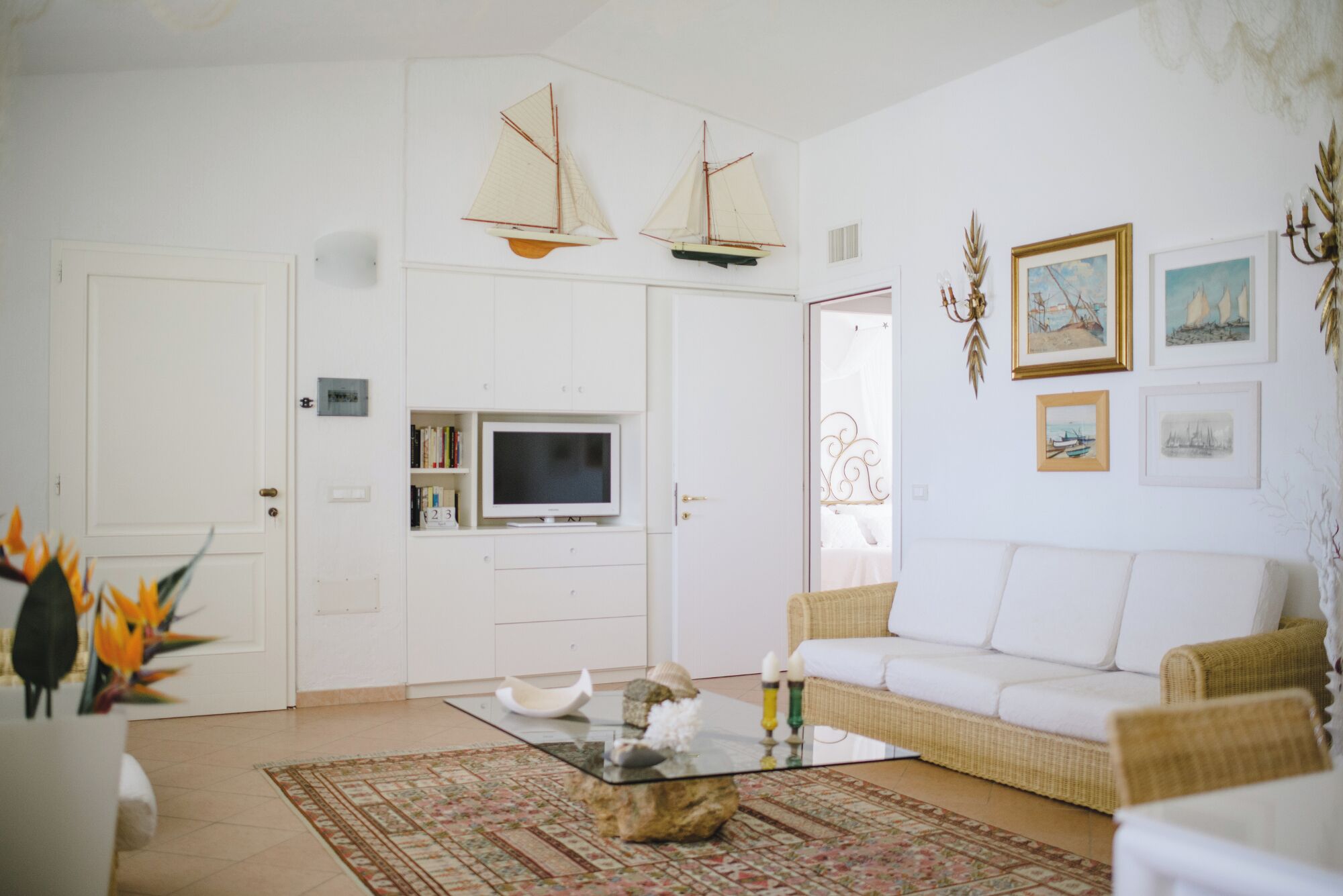 Un salon avec un canapé blanc, une table basse en verre et un téléviseur. Deux maquettes de voiliers sont accrochées au mur, et une porte porte une plaque avec un numéro.