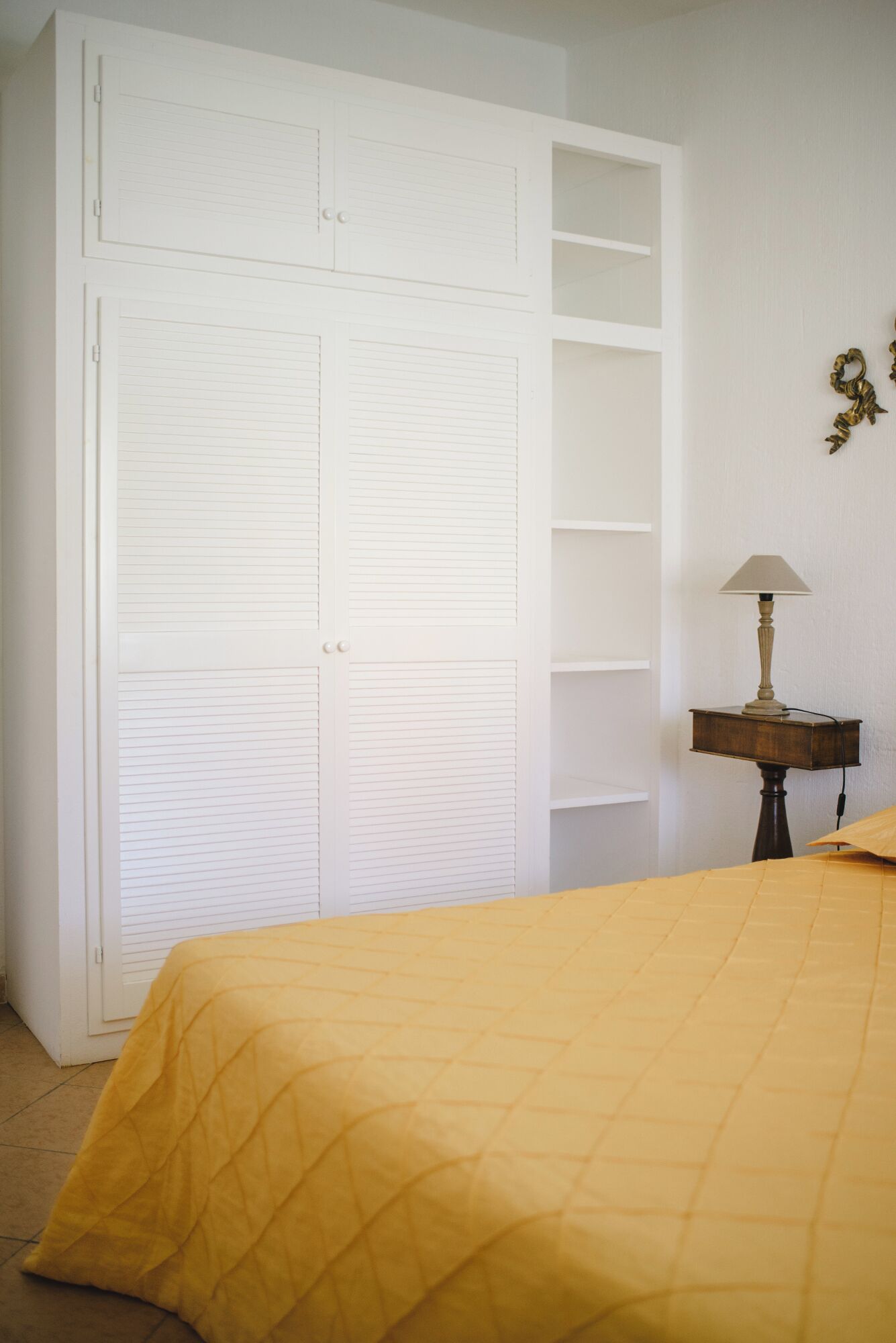 A bedroom with a large white wardrobe, a bedside table with a lamp, and a yellow bedspread.
