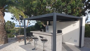 Zona de bar al aire libre con mostrador de piedra, tres taburetes y estructura de techo metálica.