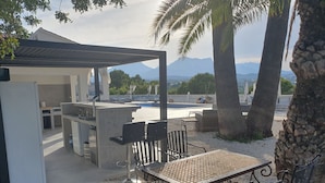 Un área de cocina al aire libre con vista a la piscina, una mesa de comedor con sillas y una estructura de pérgola.
