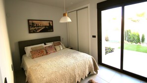 A bedroom with a large bed, a hanging light fixture, a painting on the wall, and a sliding glass door leading to a garden. - New villa with private pool (orihuela costa)