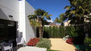A modern outdoor patio with a white wall, two white chairs, a small table, and a variety of plants including palm trees and red flowers. - New villa with private pool (orihuela costa)
