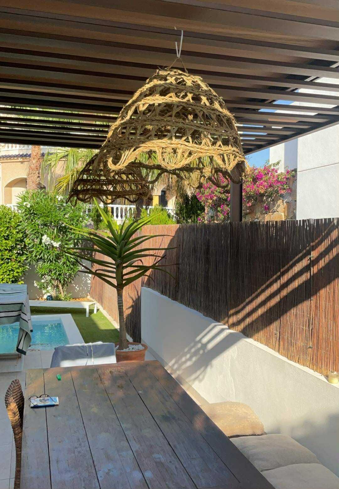 A covered outdoor seating area with a wooden table, a hanging decorative structure, and a small pool in the background.