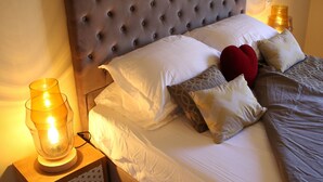 Un lit bien fait, avec une tête de lit capitonnée, une literie blanche et des coussins décoratifs, dont un coussin rouge en forme de cœur.