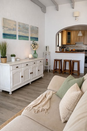 A living room with a sofa, a white cabinet, and a kitchen area with wooden cabinets and a bar stool.