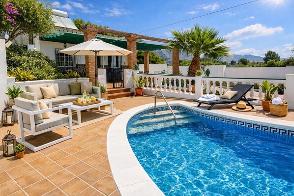 A pool area with a white outdoor sofa, a sun lounger, and a pergola with a white umbrella.