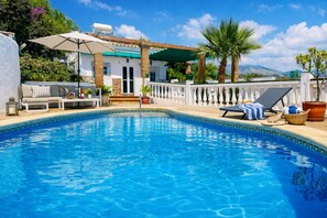 A swimming pool with lounge chairs, a covered seating area, and a view of mountains.