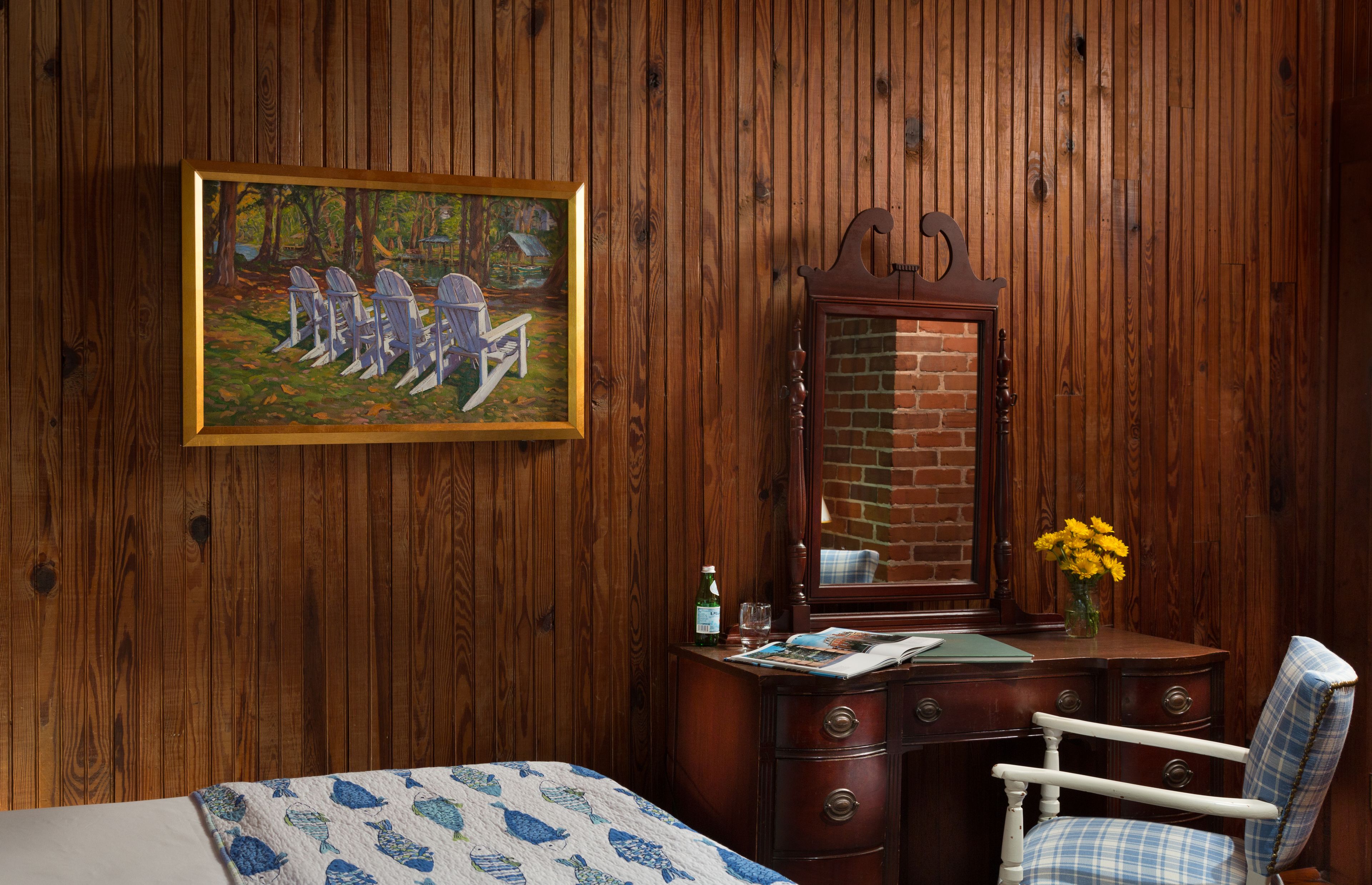 A wooden-paneled room with a bed, a dresser, a chair, a painting, and a mirror.