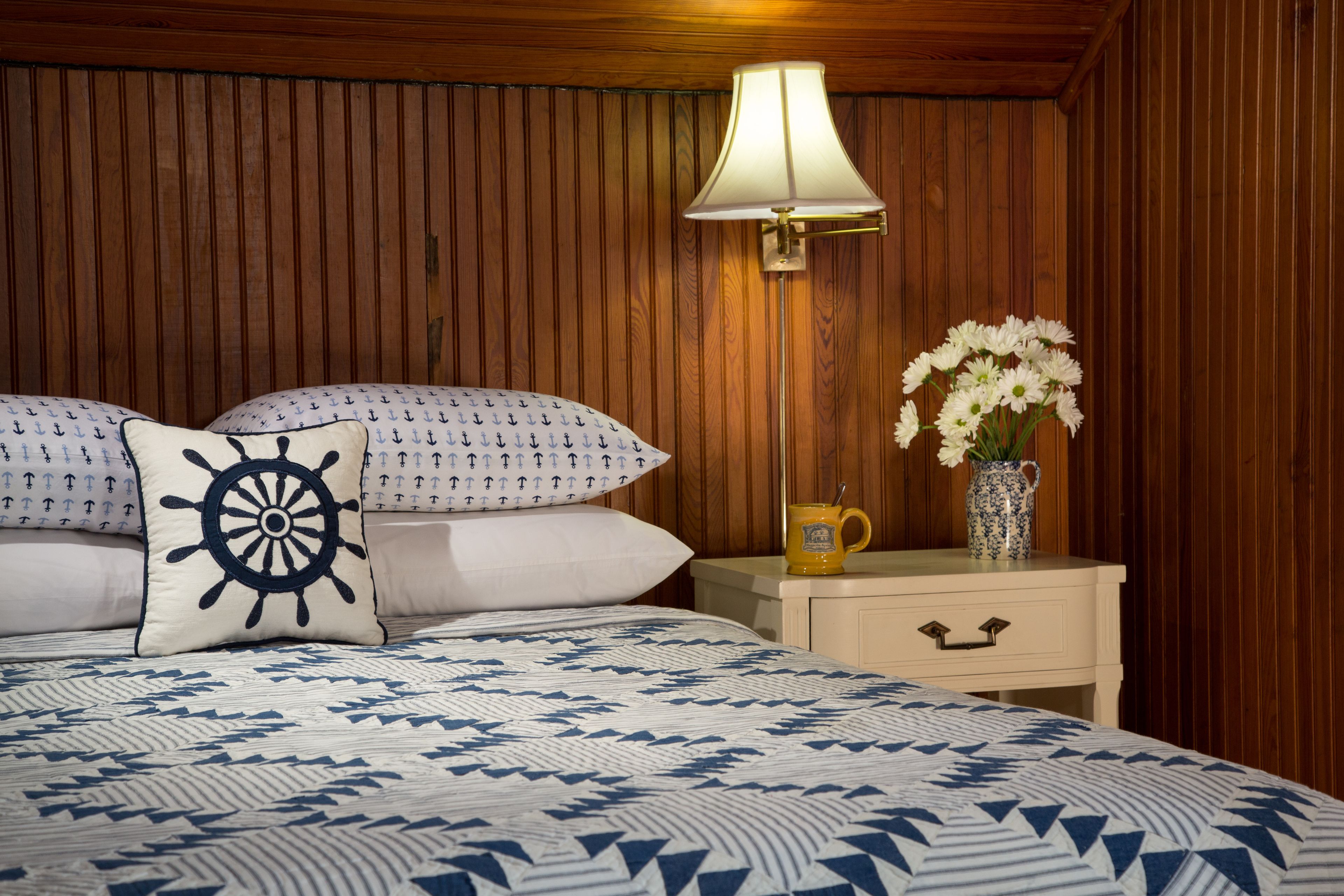 Un dormitorio con pared revestida de madera, una cama con colcha azul y blanca, una almohada con forma de timón de barco, una mesita de noche con una lámpara, y un jarrón con flores.