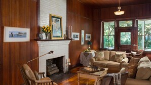 A cozy living room with a fireplace, wooden walls, and a large window offering a view of the outdoors. - Magnolia Springs B&B (Magnolia Springs)