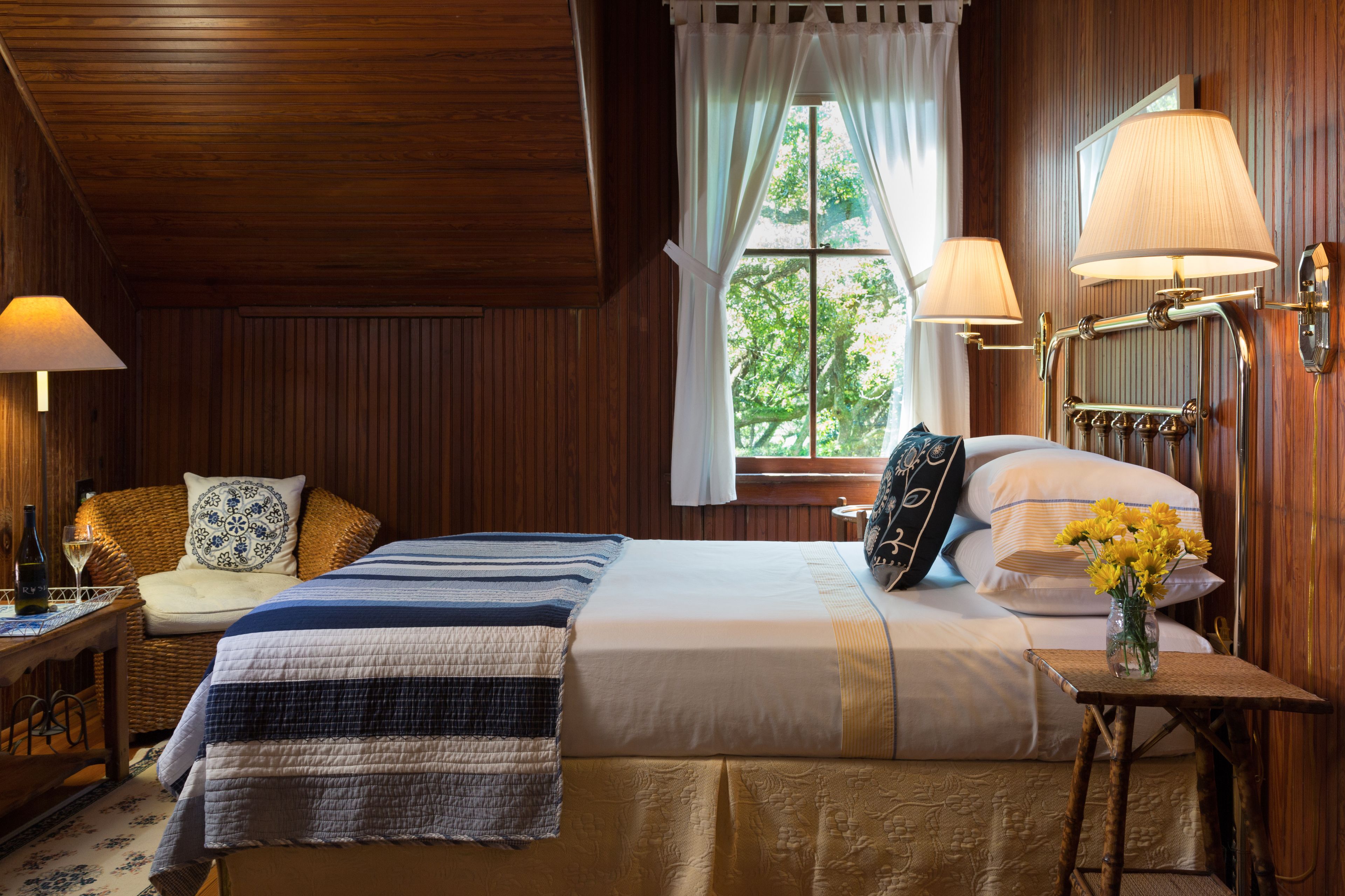 A wooden-paneled room with a bed, two bedside lamps, a window with curtains, and a small table with a vase of flowers.