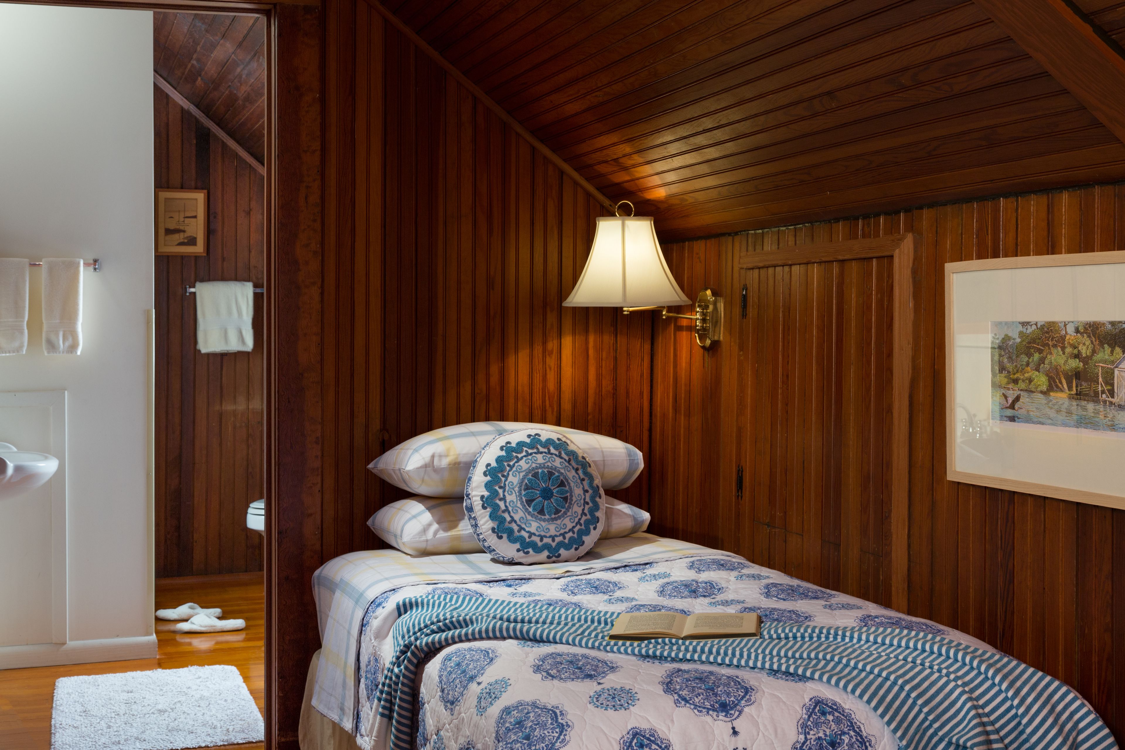 Une chambre avec des lambris en bois, un lit recouvert d’une courtepointe à motifs bleus et blancs, une lampe blanche et un tableau encadré accroché au mur.