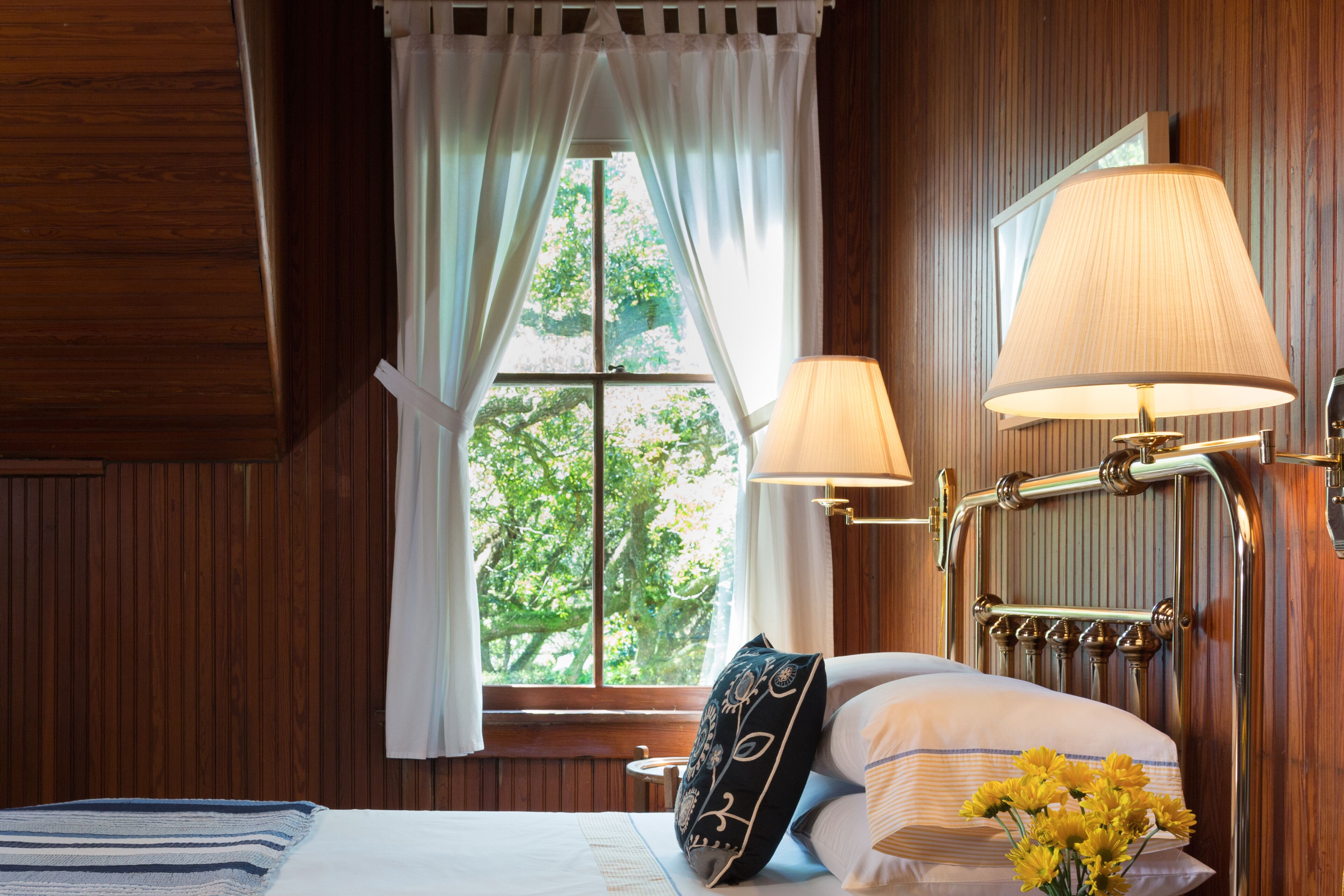 Une pièce aux murs lambrissés, avec un lit, une fenêtre avec des rideaux, une lampe et une petite table sur laquelle est posé un vase de fleurs.